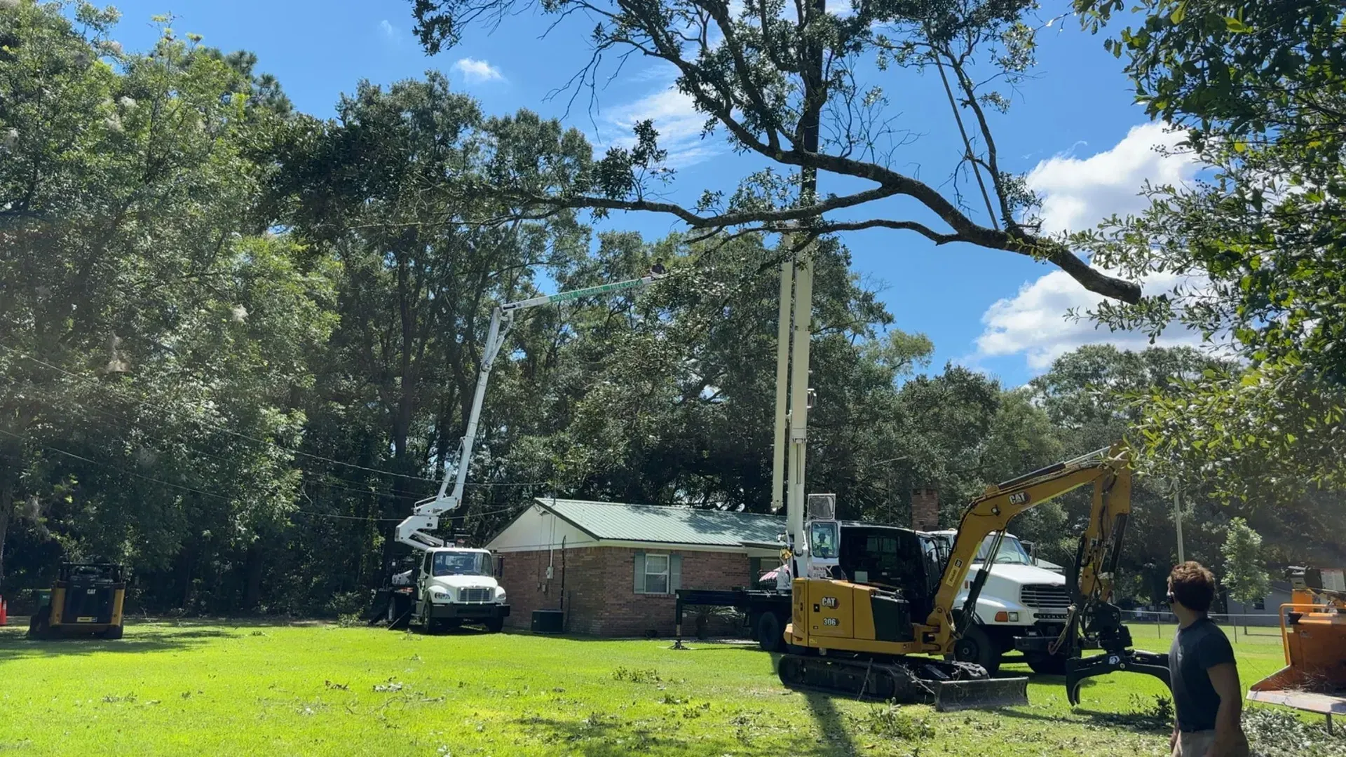 Tree removal: Crew using machinery to remove branches over a house; sunny day with green yard and trees.