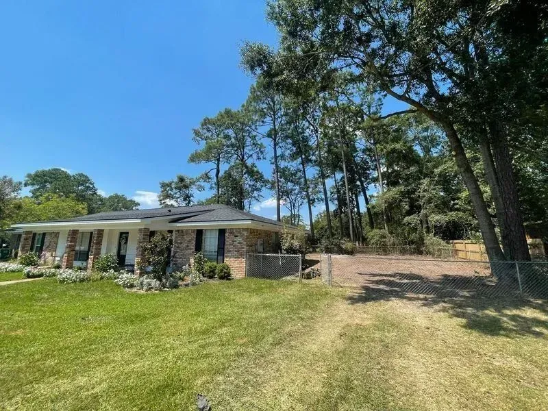 A single-story brick house with a dark roof and a grassy yard. Tall trees line the driveway.