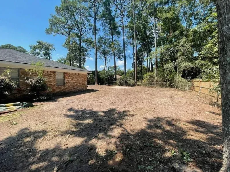 Cleared backyard with brown ground, trees, and a brick house in the background under a blue sky.