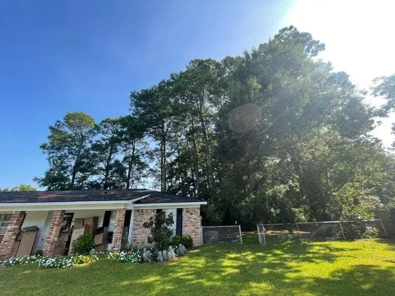 A residential house with a brick exterior is next to tall trees under a bright blue sky.