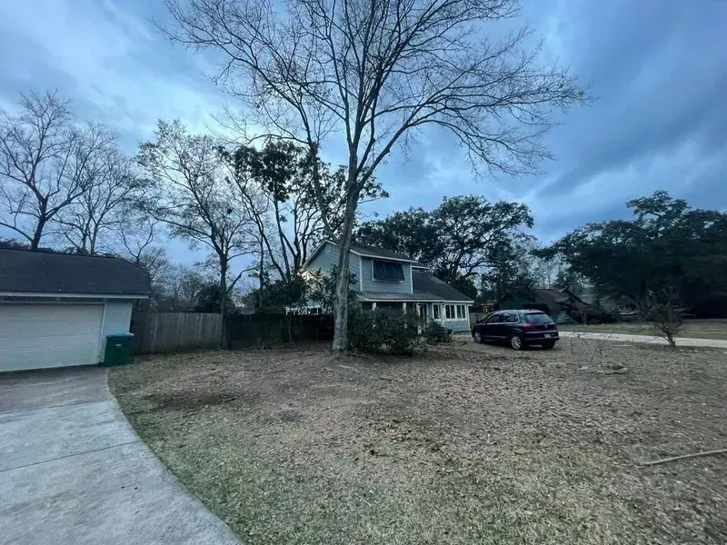 House with large tree in front yard, gray car parked on the right, cloudy sky.