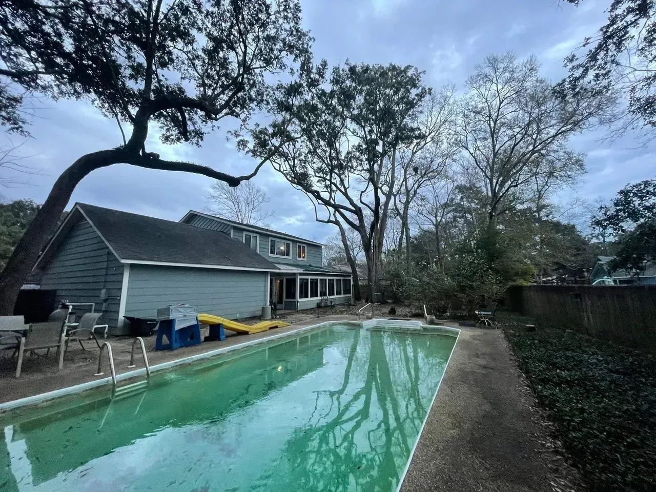 Backyard with a green pool, light blue house, and trees under an overcast sky.