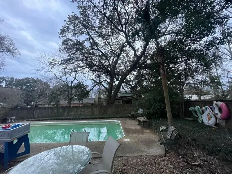 A backyard with a pool, trees, and a cloudy sky.  Poolside seating and a table are visible.