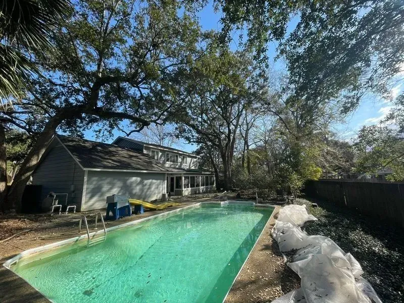 Backyard with a swimming pool, house, and large trees on a sunny day.