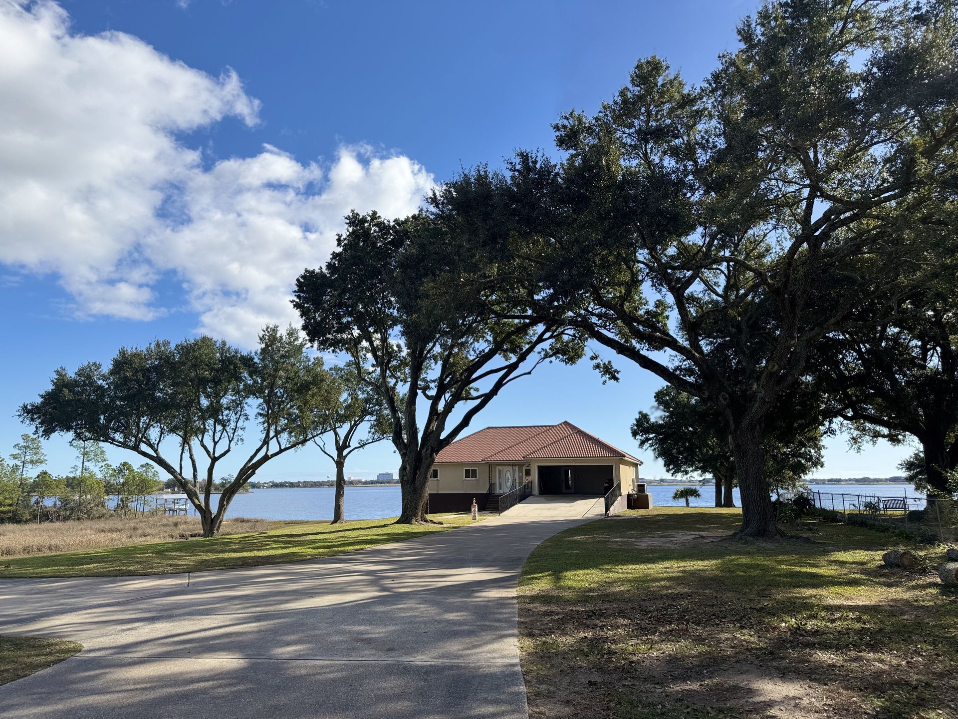 Driveway leads to a house with a brown roof, framed by large trees against a blue sky over water.