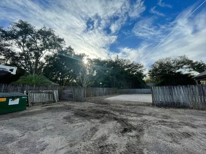 Gravel lot with weathered wooden fence, green dumpster, and trees under a blue sky with streaky clouds.