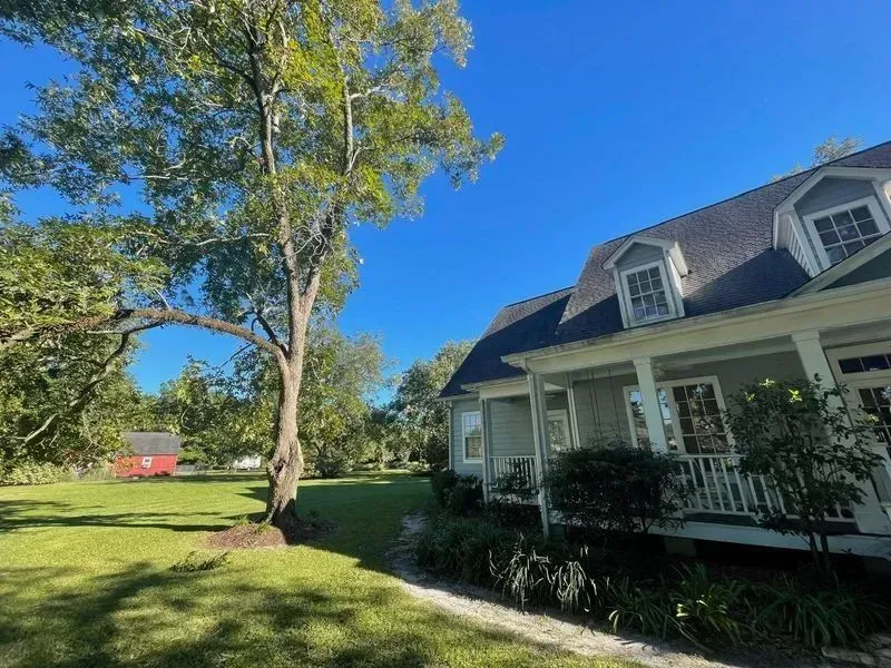 House with porch, tree, and lawn on a sunny day; small red building in the distance.