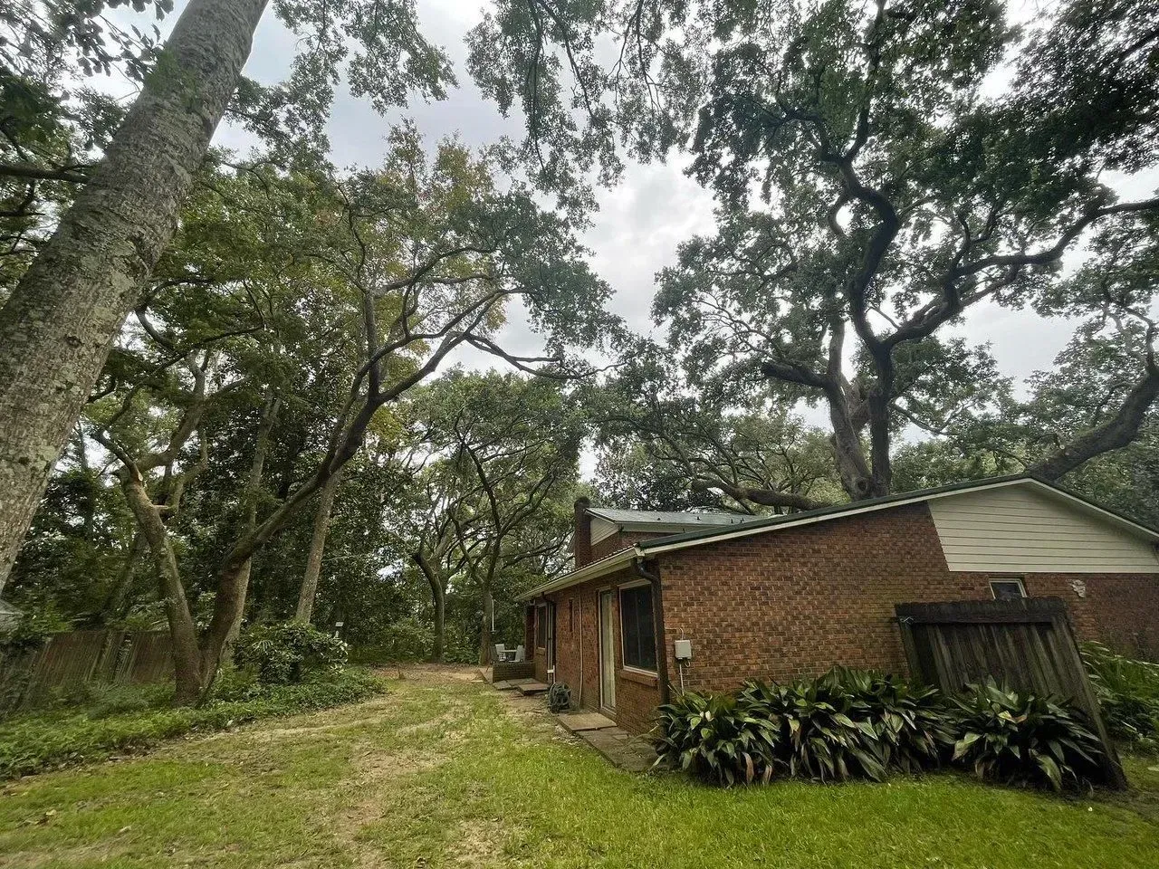 Brick building with a wooden door, surrounded by lush green trees and foliage, under a cloudy sky.