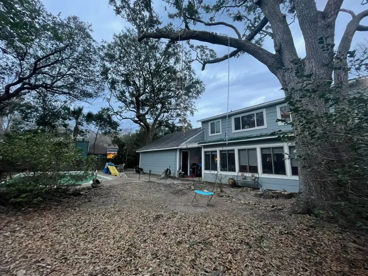 Backyard of a two-story blue house under a cloudy sky, with trees and fallen leaves covering the ground.