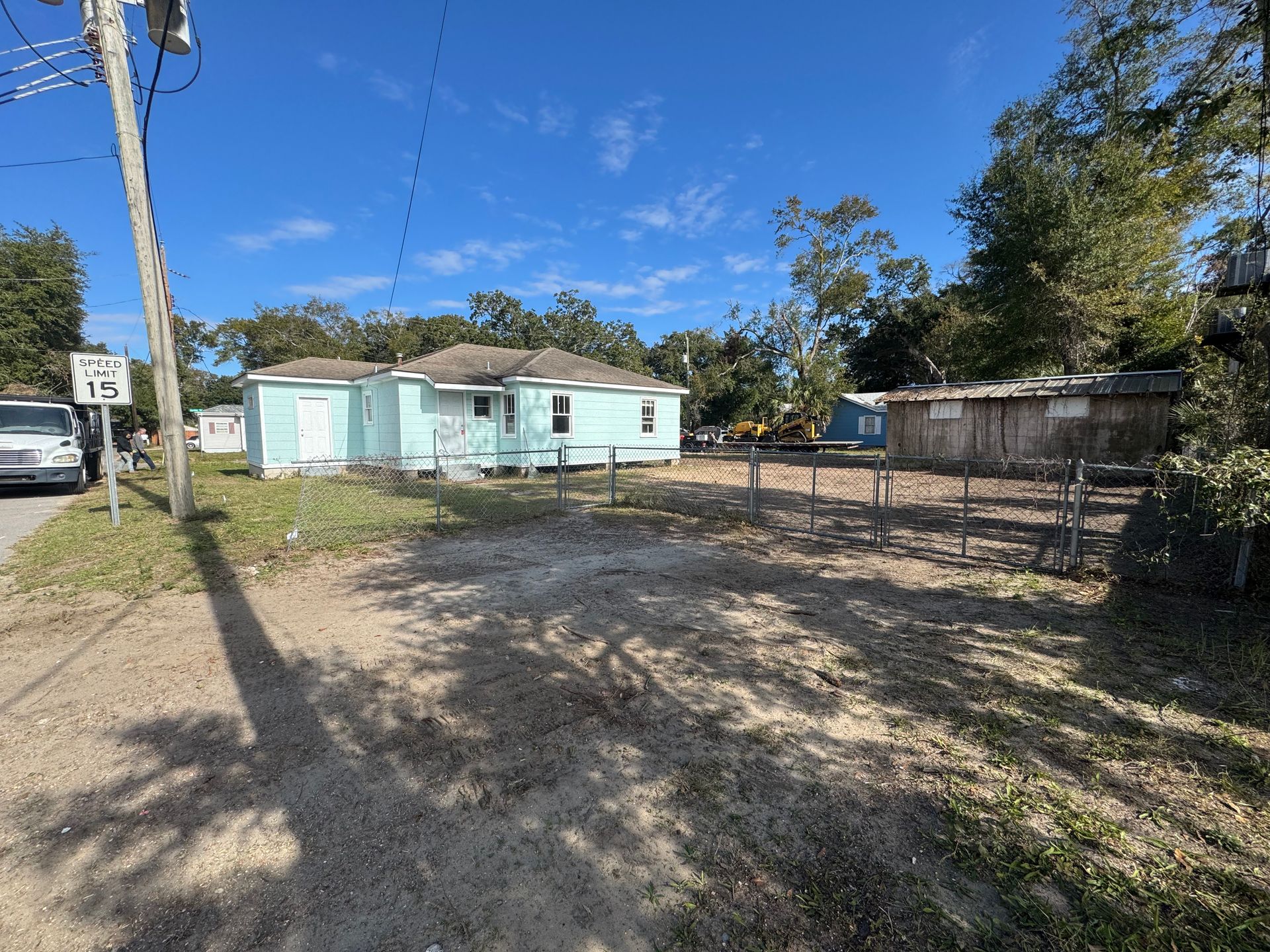 A pale blue house with a fence, dirt yard, and telephone pole on a sunny day.