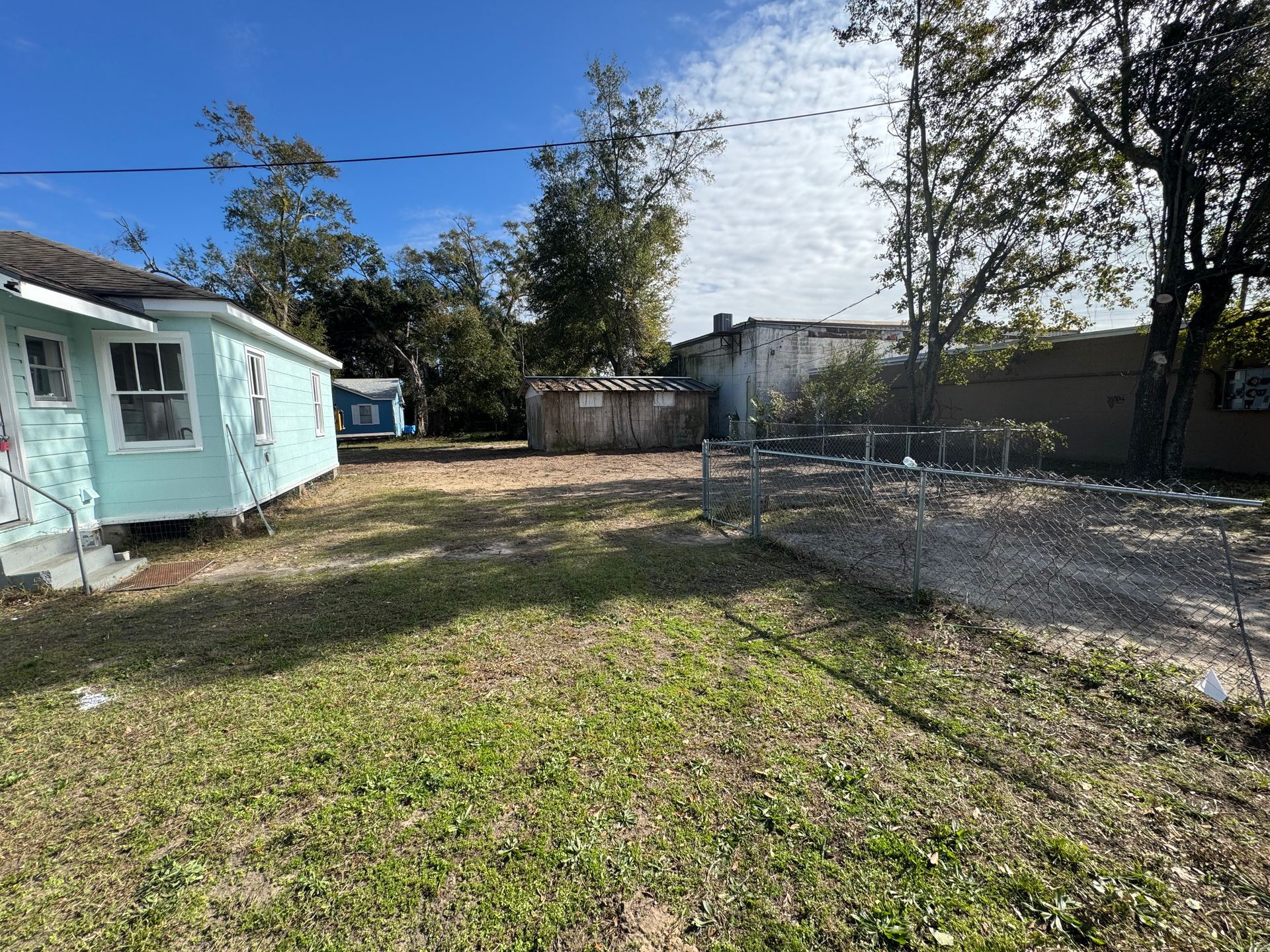 Backyard with a light blue house, trees, and brown shed under a blue sky.