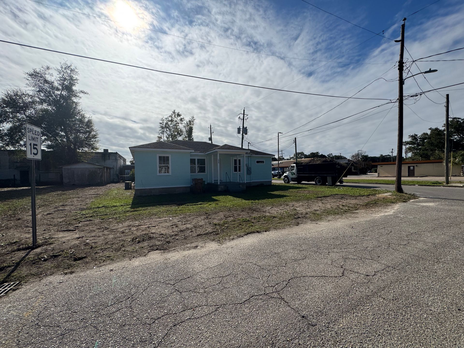 A small, light blue house with a grassy front yard, near a road under a sunny, cloudy sky.