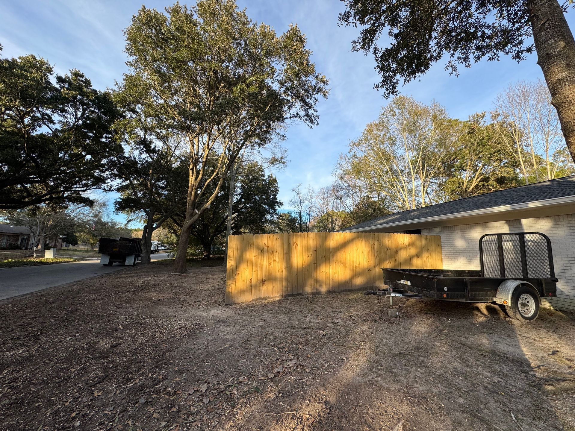 A sunny daytime view of a yard with a new wooden fence, trailer, and trees.
