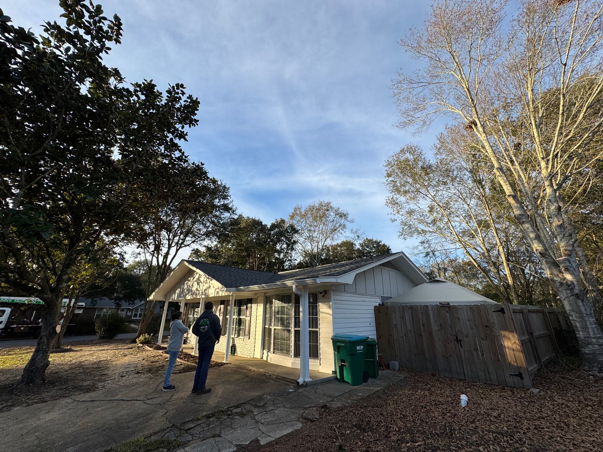 Two people standing near a house with visible roof damage, debris, and trees on a sunny day.