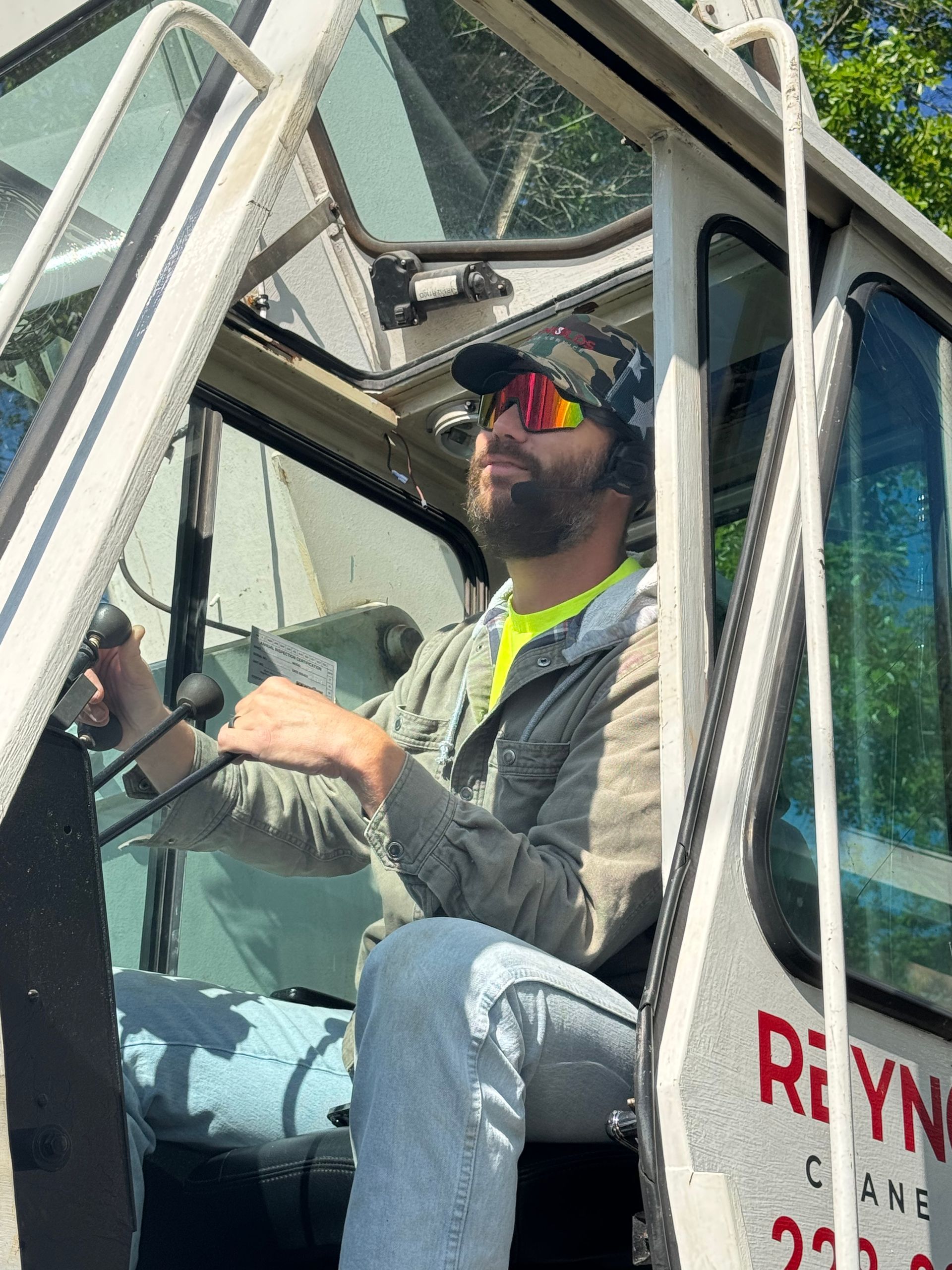 Man operating crane, wearing sunglasses and a camo hat, in a white cab with 