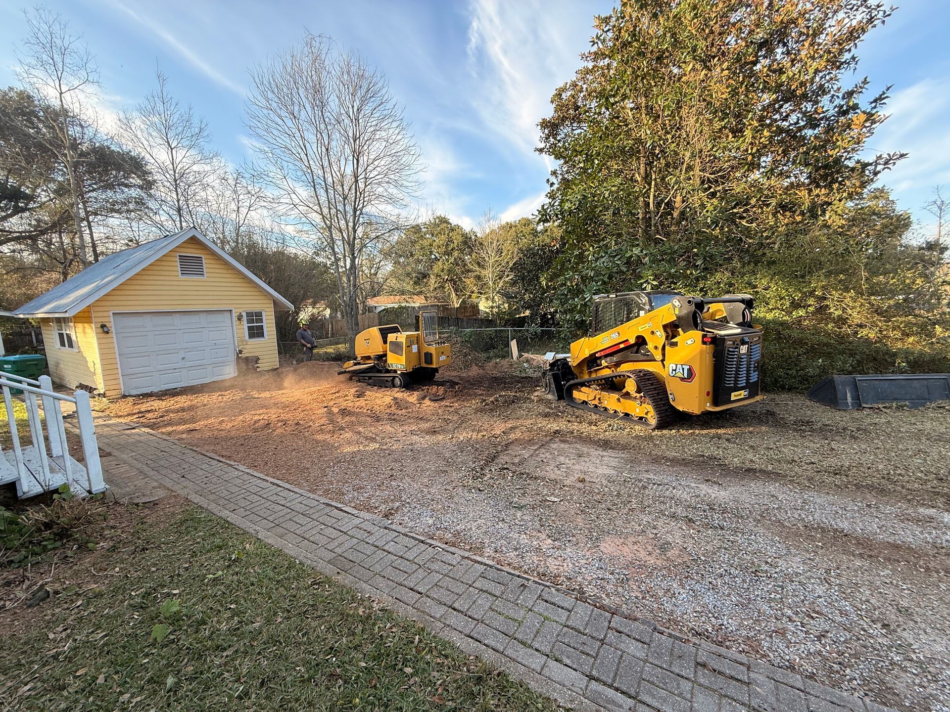 Construction site with two yellow skid steers, garage, and gravel driveway.