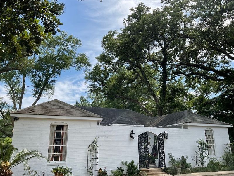 White brick house with dark roof, ornate gate, and lush green trees against a blue sky.