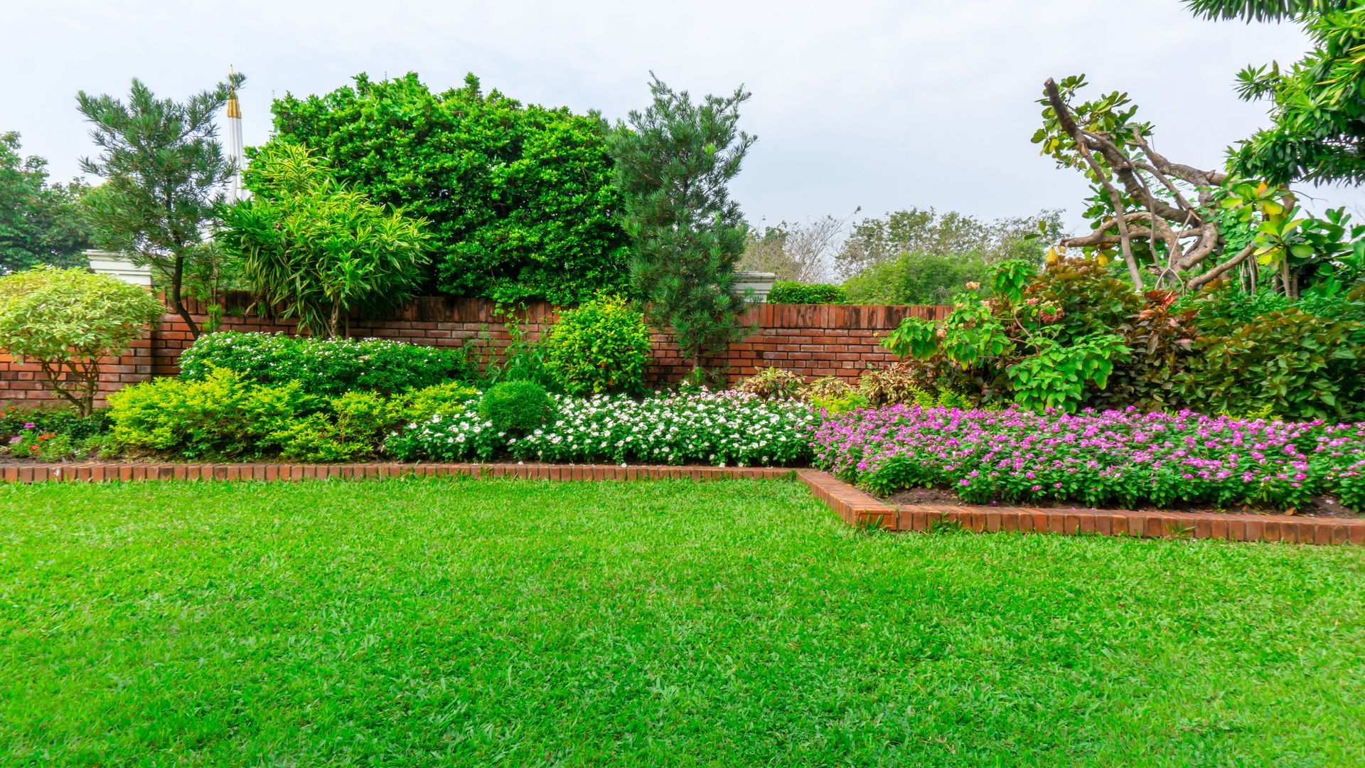 Green lawn with a brick-lined garden bed filled with colorful flowers and shrubs under a cloudy sky.