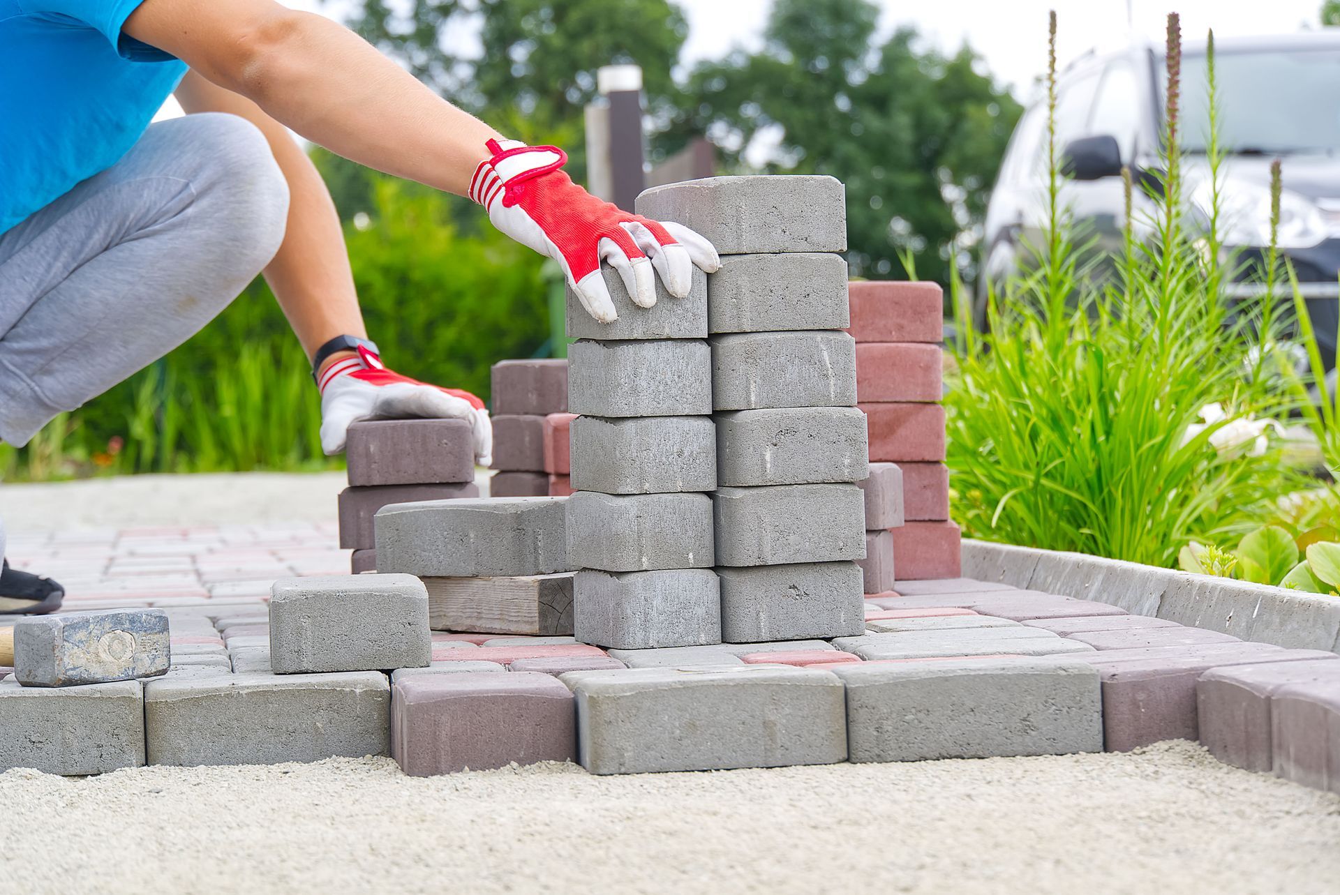 Person laying gray and red paving stones, wearing gloves, outdoors.