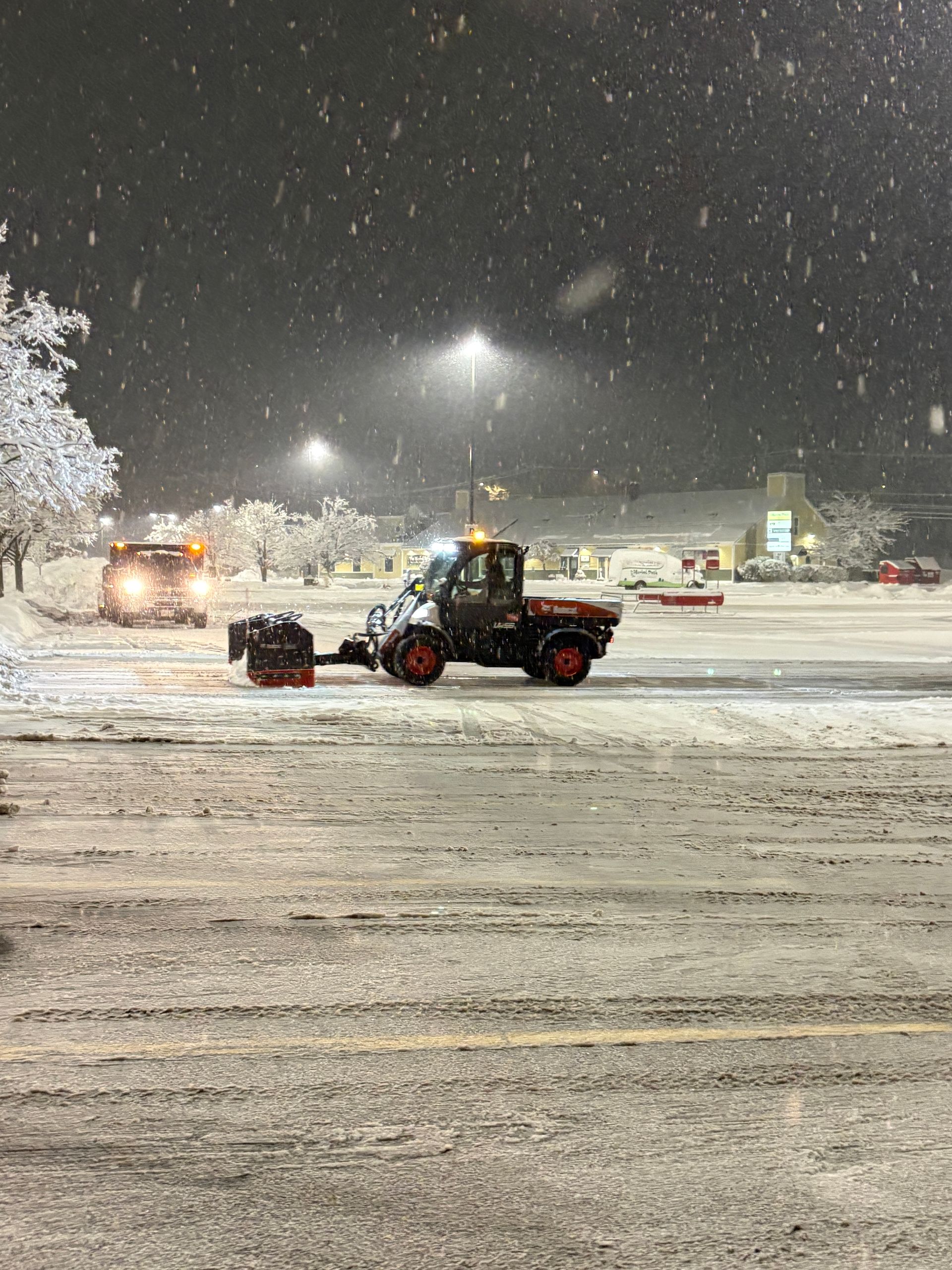 Snow plows clearing a snowy parking lot at night. Trees and lights visible.