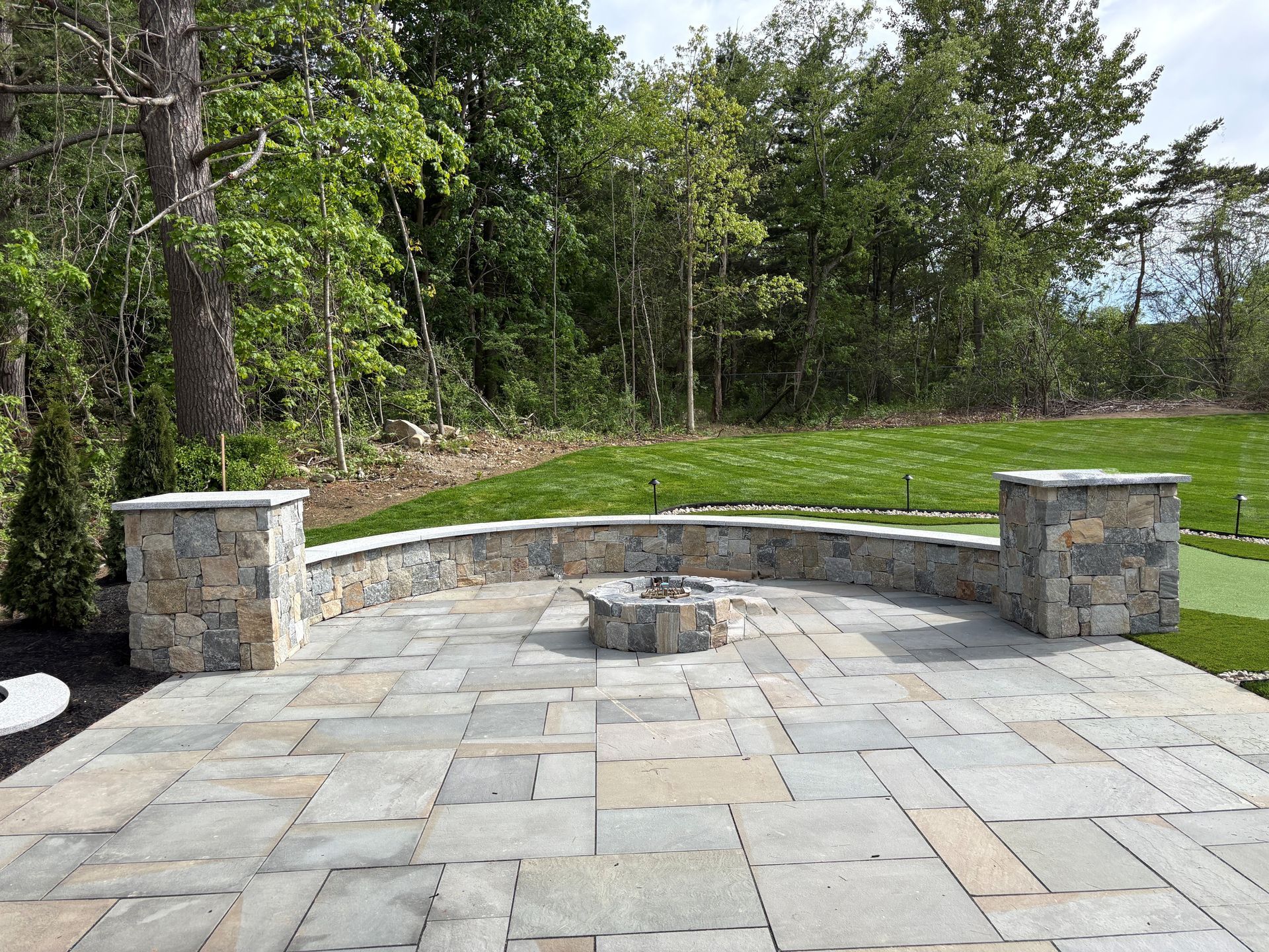 Stone patio with a curved bench and fire pit, bordered by trees and green lawn.