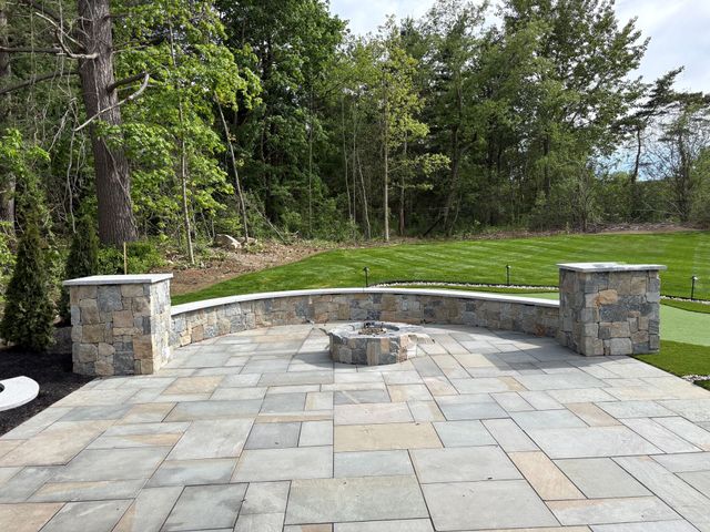 Stone patio with a curved bench and fire pit, bordered by trees and green lawn.