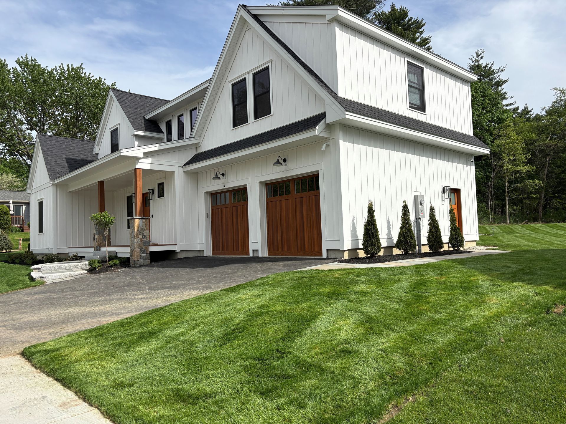 White two-story house with a front garage, sloped roof, and manicured green lawn on a wooded lot