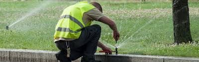 Worker in a neon safety vest watering grass beside a sidewalk with a hose