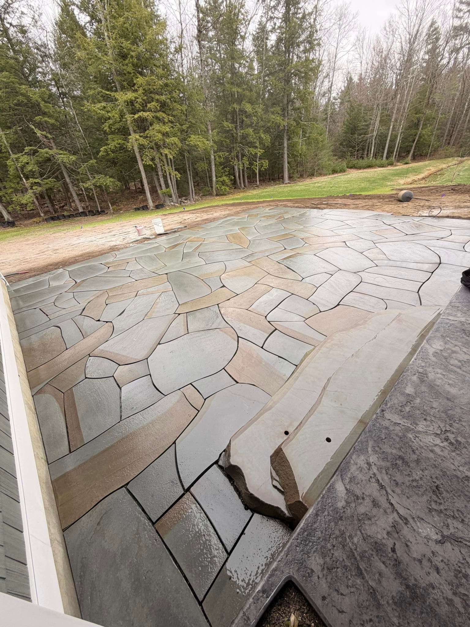 Broken stone patio beside a forested yard, with a cracked slab and a gravel edge.