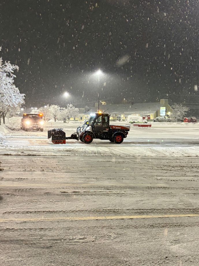 Snow plows clearing a snowy parking lot at night. Trees and lights visible.