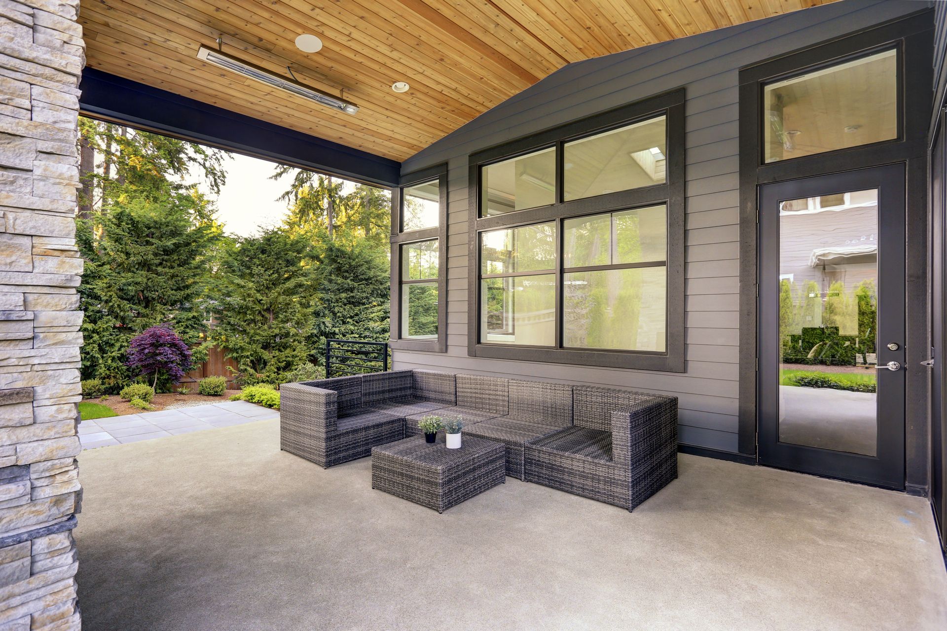 Covered patio with gray wicker furniture, overlooking a garden. Wooden ceiling, gray siding, and a stone wall.