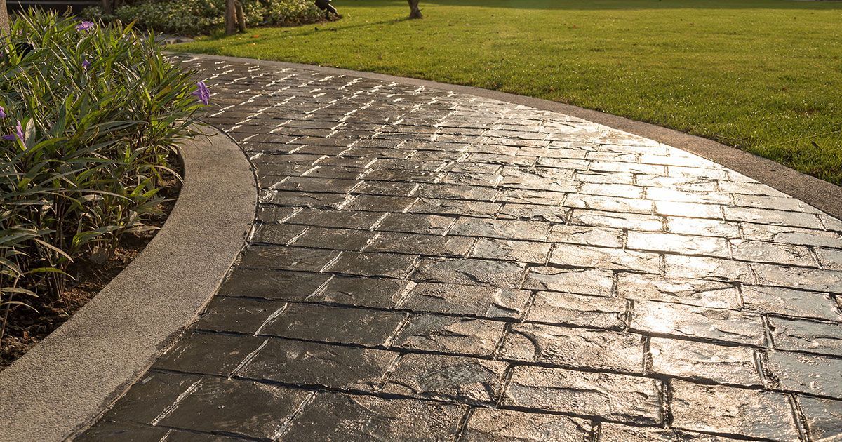 Brick-patterned concrete pathway curves through a green lawn, edged by a textured border and plants.