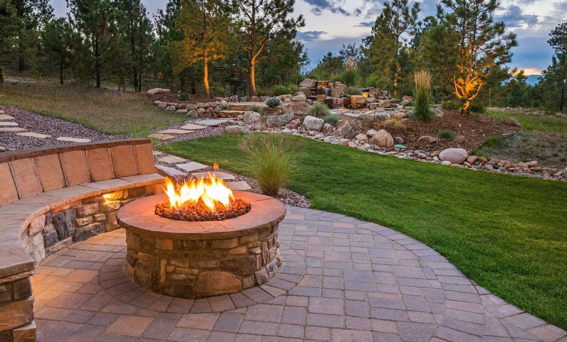 Stone fire pit with flames, curved bench, paved patio, green lawn, trees in the background.