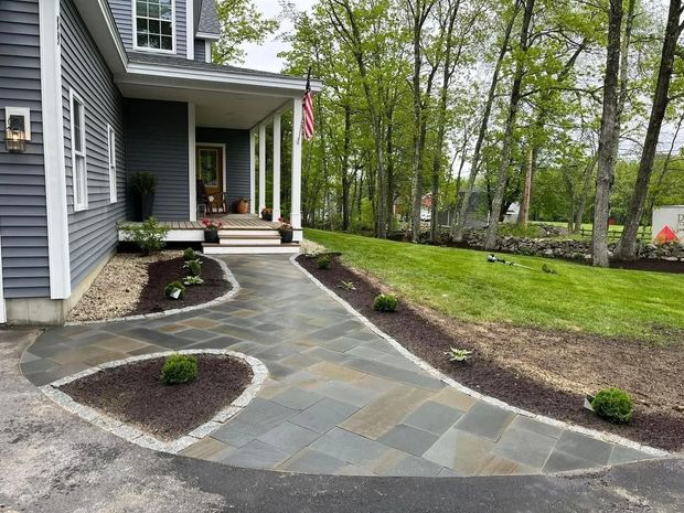 A paved walkway leads to a house with a porch, bordered by landscaping and a lawn.