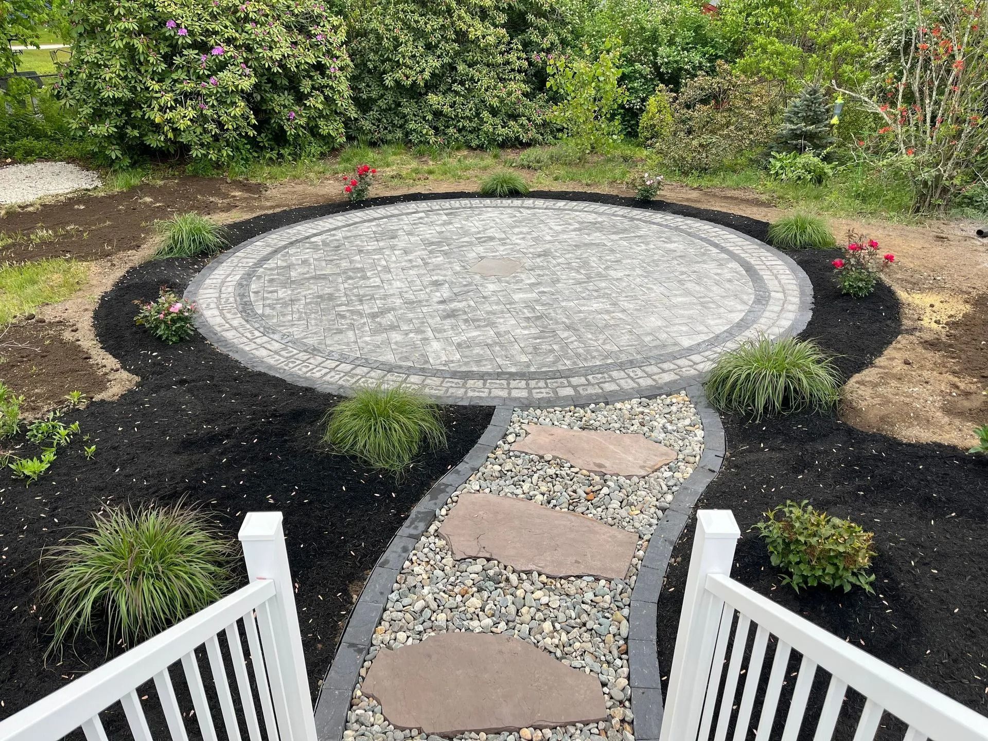 Circular stone patio with stepping stone path, surrounded by mulch and greenery.