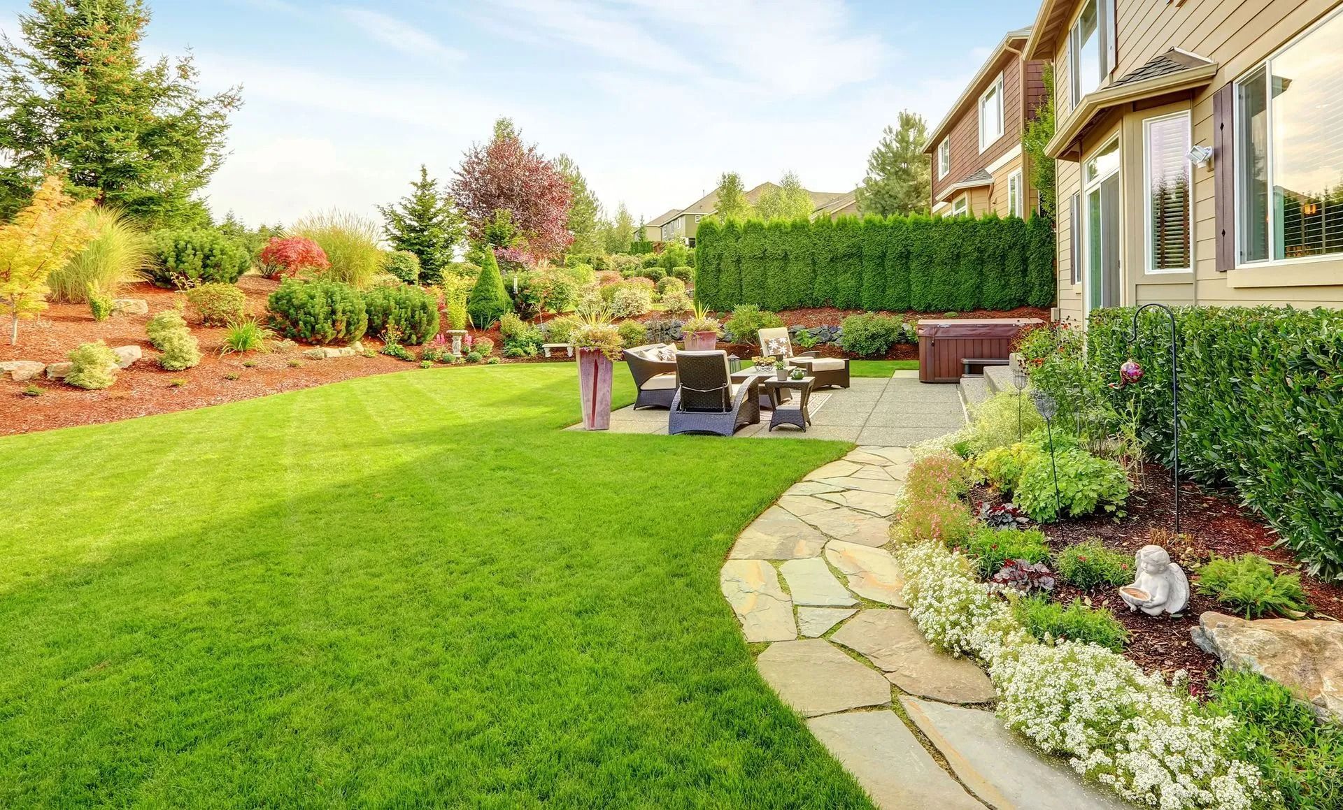 Lush green backyard with lawn, stone path, patio furniture, and surrounding flowerbeds.