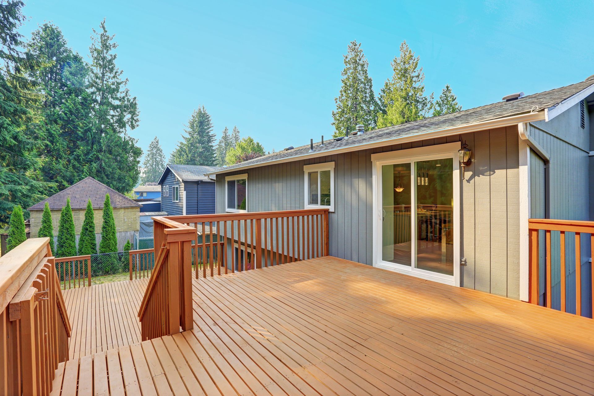 Wooden backyard deck beside a gray house, with trees and a blue sky in the background