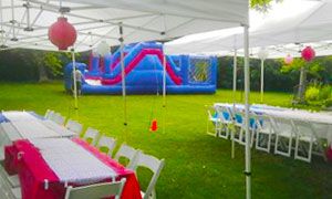 Outdoor party tent with tables and chairs on a lawn, a red lantern, and a colorful inflatable bounce house in back