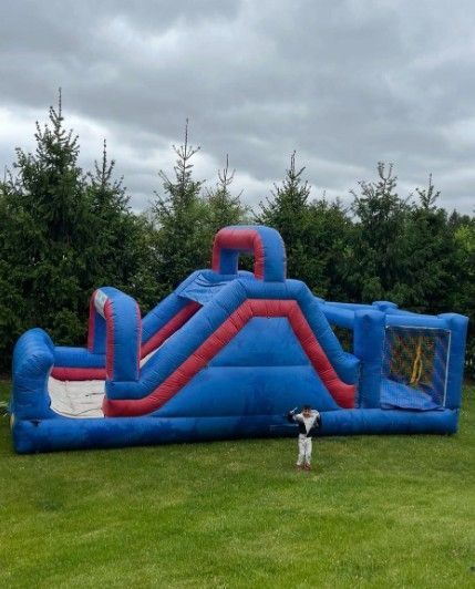 Blue and red inflatable obstacle course on a lawn, with a dog standing in front and cloudy sky behind.