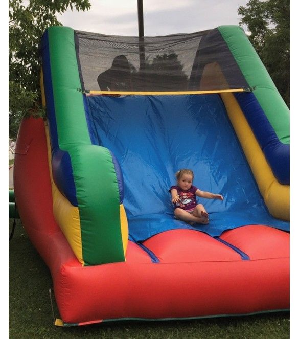 A child slides down a colorful, multi-colored inflatable slide outdoors on a patch of grass.