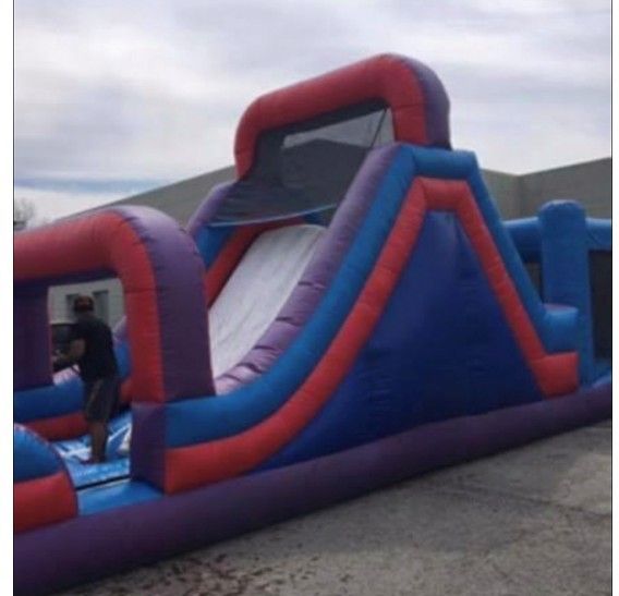 A large, red, blue, and purple inflatable obstacle course with a central slide, set up on an outdoor paved surface.