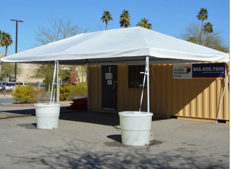 White canopy tent outside a tan container office in a parking lot, with palm trees in the background.