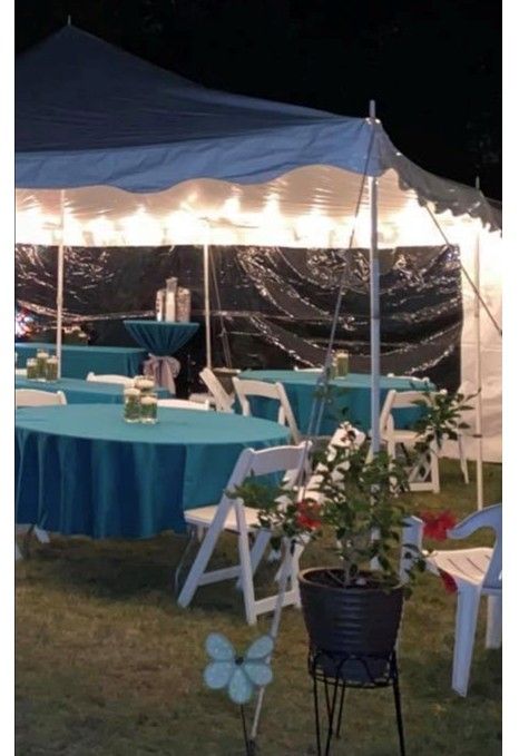 Lit tent at night with teal-covered tables, white chairs, and string lights at an outdoor event.