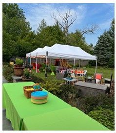 Backyard party setup with white tents, green tablecloth, and colorful baskets amid garden greenery.