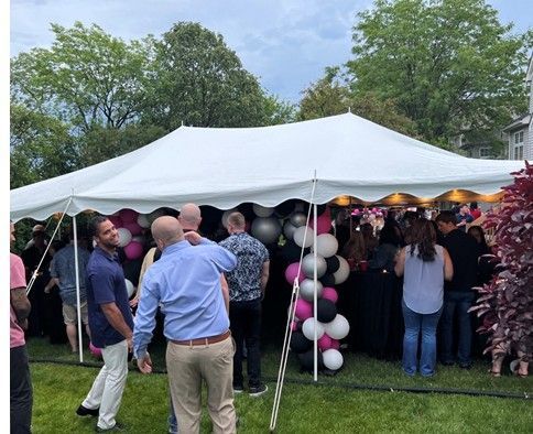 People gathered under a white event tent on a grassy lawn, with pink-and-white balloon decorations.