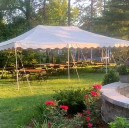 White outdoor tent with picnic tables in a grassy yard, with pink flowers in the foreground and trees behind.