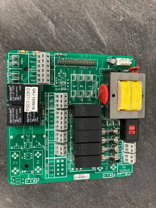 Green electronic control circuit board with relays, connectors, and a yellow coil on a gray surface