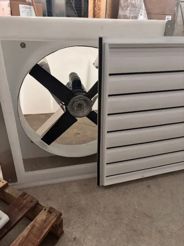 Large square ventilation fan with black blades and white slatted cover in a workshop.