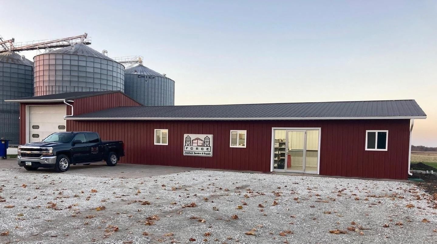 Maroon metal building with a black pickup parked outside beside grain silos on a gravel lot.