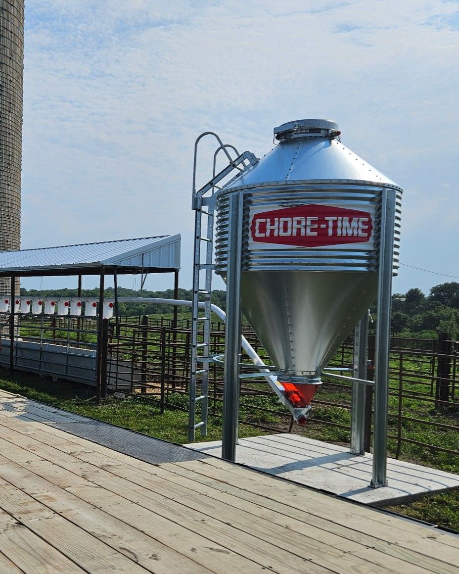 Large silver grain silo labeled “CHOICE-TIME” beside a wooden boardwalk and fenced farm field